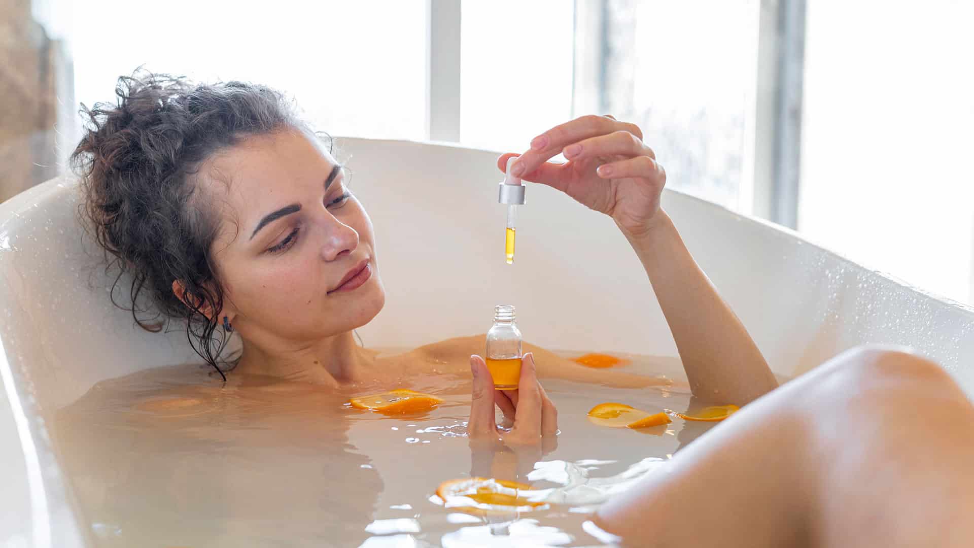 een vrouw die in een badkuip zit en een fles vloeistof vasthoudt.